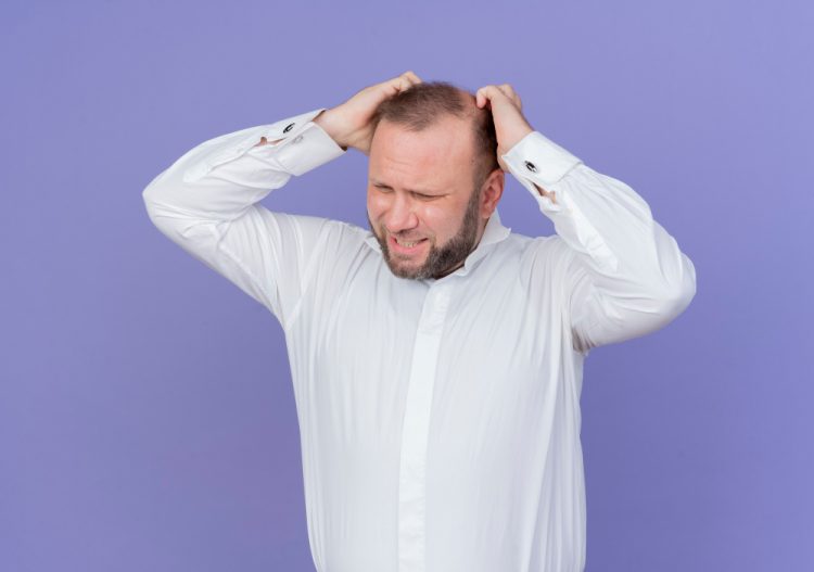 upset bearded man wearing white shirt touching his head being frustrated standing blue wall