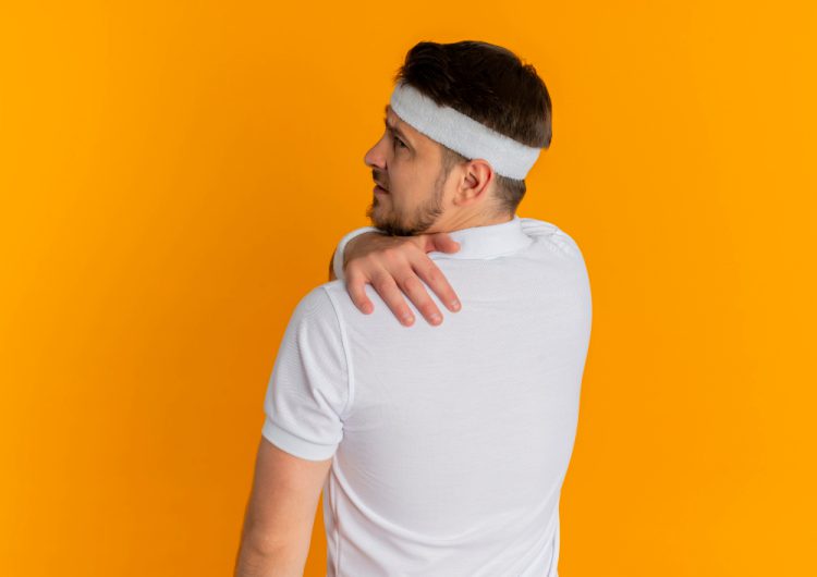 young fitness man white shirt with headband standing with his back touching his shoulder having pain orange wall